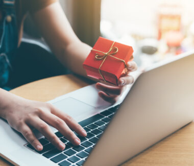 Woman at her laptop holding a red gift box as she shops online.